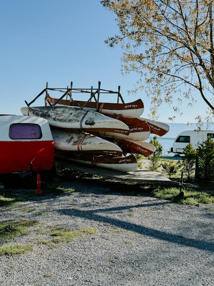 A collection of canoes stacked near a red camper trailer at a scenic lakeside campsite.